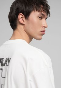 T-shirt in cotone bianco con scollo rotondo e maniche corte. Presenta una stampa di testo nero audace sul retro, con una vestibilità leggermente oversize.