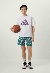 Henkilö, joka pitää käsissään koripalloa, pukeutunut valkoiseen Adidas-paitaan, tealsävyisiin kuvioituihin shortseihin, valkoisiin sukkiin sekä mustavalkoisiin tennareihin, seisomassa yksinkertaisella taustalla.