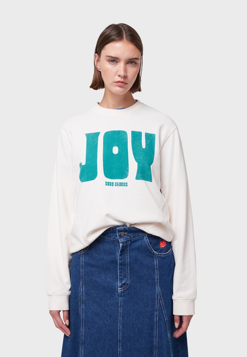 Felpa bianca con grande testo e logo "JOY" in teal, abbinata a una gonna di jeans con un dettaglio a cuore rosso. Vestibilità semplice e rilassata.