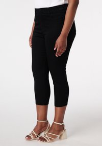 Pantalons capri noirs avec une taille élastique, présentant une coupe ajustée et une texture en denim douce. Portés avec des sandales à talons carrés beiges.