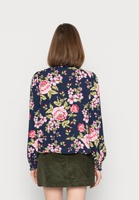 Blusa floral com fundo azul-marinho, featuring grandes rosas cor-de-rosa e vermelhas, folhas verdes, mangas longas e um corte descontraído. Acima de uma saia de veludo cotelê verde.