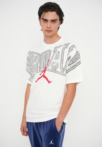 Valkoinen puuvillapaita, jossa on musta ja punainen graafinen suunnittelu, jossa on teksti "JORDAN" ja punainen siluetti pelaajasta. Lyhyet hihat.