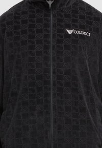Veste zippée en velours noir avec un motif géométrique subtil et un logo Colucci brodé en blanc avec des ailes sur la poitrine.