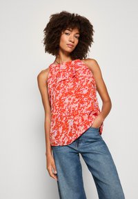 Haut sans manches rouge et rose avec un motif, doté d'un col rond et de détails froncés à l'avant, associé à un jean bleu. Texture de tissu lisse.