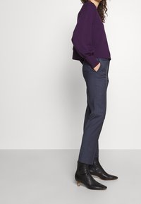 Pull cropped violet, pantalon bleu marine ajusté et bottes noires à talons avec accents dorés. Texture lisse, design ajusté.