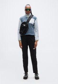 Φλις πουλόβερ σε ανοιχτό γαλάζιο με μαύρες λεπτομέρειες, μαύρο παντελόνι καμπαρντίνα τύπου cargo και μαύρη τσάντα χιαστί. Διαυγή, αντανακλαστικά γυαλιά ηλίου και παπούτσια με υφή.