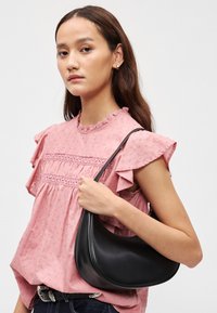 Crna kožna torba hobo s zakrivljenim oblikom, glatkom teksturom i jednim remenom, uparen s ružičastom bluzom s volanima koja ima cvjetne uzorke.