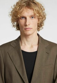 Olivzaļš žakete ar piemērotu piegriezumu, iegriezuma atloki un vienu krūšu kabatu, valkāta pār melnu ribota auduma T-kreklu. Teksturēts audums.