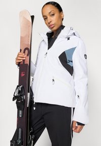 Ski jakna u bijeloj boji s crnim i plavim detaljima, vodootporni materijal, džepovi sa patentnim zatvaračem, ukrašena logotipima, uparena sa skijama i vezovima.