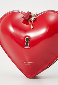 Heart-shaped red patent leather bag, featuring a gold key zipper pull, a black keyhole accent, and gold "kate spade NEW YORK" logo detail.