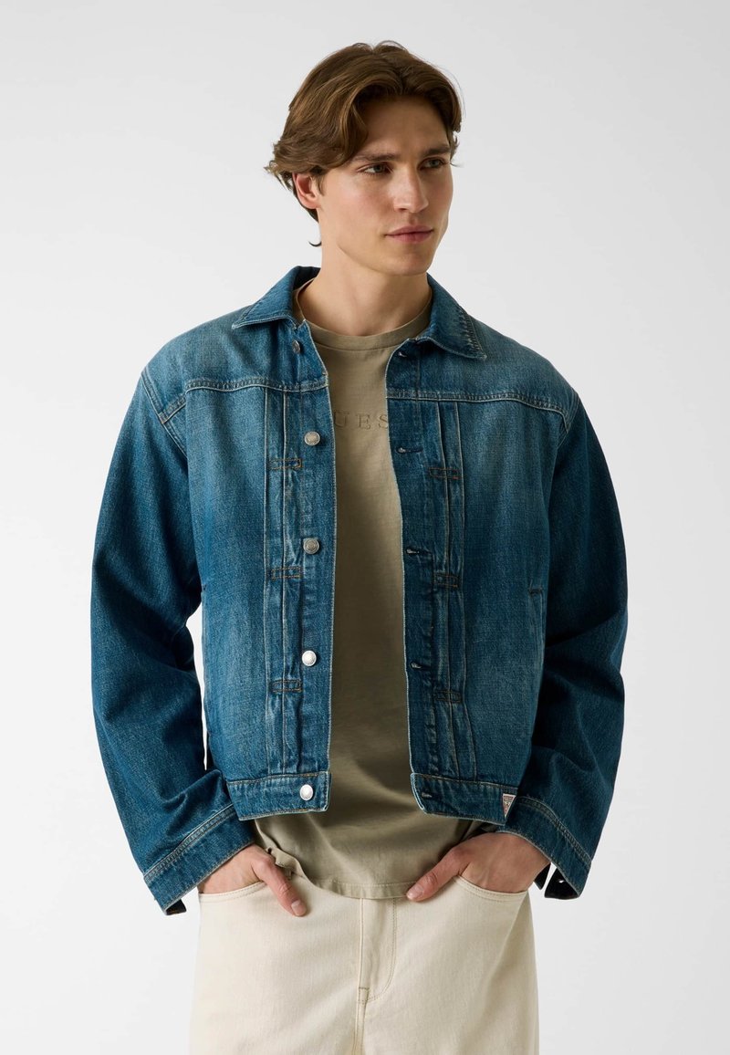 Jeune homme aux cheveux bruns portant une veste en jean bleue par-dessus un t-shirt beige et un pantalon clair, les mains dans les poches, regardant vers la droite.