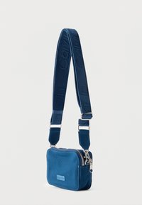 Συμπαγής μπλε υφασμάτινη τσάντα χιαστί με μπροστινό έμβλημα λογοτύπου, ασημένια φερμουάρ και φαρδιά ρυθμιζόμενη λουρίδα με ανάγλυφο το όνομα της μάρκας.