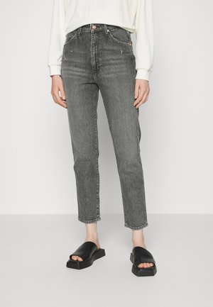 Jeans de mezclilla gris con cintura alta, corte recto y textura ligeramente desgastada. Presentan accesorios en tono cobre y un dobladillo sin terminar. Sandalias negras de tipo slide.