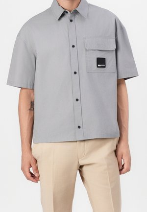 Uomo con camicia a maniche corte grigio chiaro con taschino frontale a pattina e pantaloni beige, in piedi davanti a uno sfondo semplice.