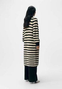 Strickjacke mit vertikalen schwarzen und cremefarbenen Streifen, langen Ärmeln und einer gerippten Textur. Reicht bis zur Mitte des Waden, kombiniert mit tief geschnittenen lässigen Hose.