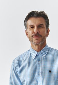 Camisa de cuadros azul y blanca con cuello button-down, que presenta un pequeño logo de oso bordado en el lado izquierdo del pecho. Material de algodón.