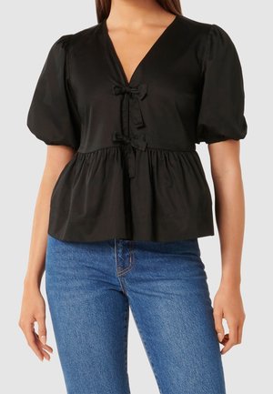 Femme portant un chemisier noir à manches bouffantes avec un col en V et deux nœuds, associé à un jean en denim bleu, debout, bras détendus.