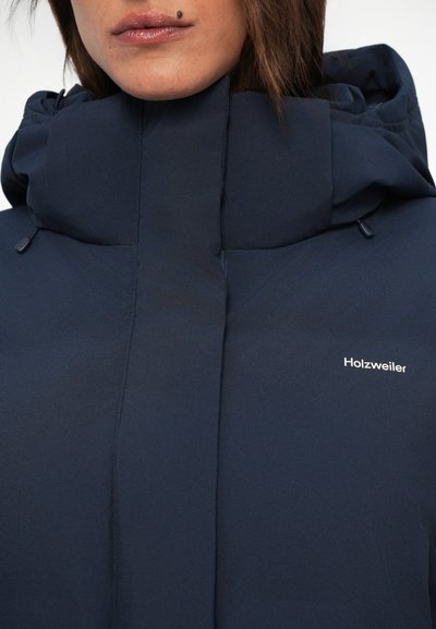 Veste à capuche bleu marine avec une finition lisse et texturée. Présente un col haut et un logo sur la poitrine. Bouts ajustables sur la capuche.