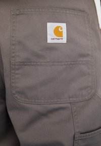 Carhartti tumedad hallid püksid, millel on ruudukujuline valge silt oranžiga logoga, ning tagaküljel nähtavad tikandite detailid tasku.