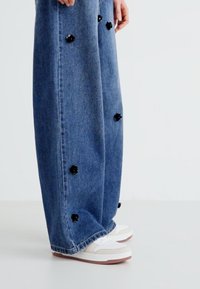 Jeans in denim a gamba larga di colore azzurro chiaro con applicazioni floreali nere. Presentano un orlo pulito e sono abbinati a sneaker bianche e beige.