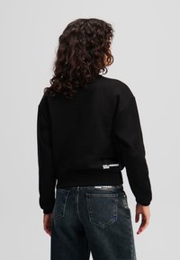 Black cropped sweatshirt with a round neckline, featuring a side pocket and logo detailing. Paired with dark denim jeans.