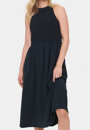 Vrouw in een mouwloze zwarte midi-jurk met een geplooide, textuurvolle lijfje en een vloeiende rok, waarbij ze lichtjes de stof aan één zijde vasthoudt.