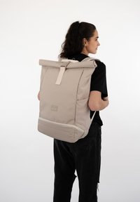 Light beige roll-top backpack with a textured fabric, adjustable straps, and a front logo patch, carried on the back.