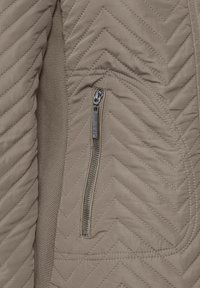 Beżowa pikowana kurtka z wzorem jodełki, wyposażona w zasuwaną boczną kieszeń i gładką fakturę tkaniny. Akcenty obejmują srebrny zamek błyskawiczny.