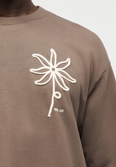 Ανοιχτό καφέ πουκάμισο με λευκή κεντημένη αφηρημένη λουλούδι και μικρό κείμενο "NN. 07" στο στήθος.