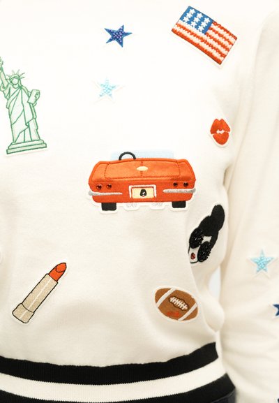 Άσπρο πουλόβερ με κεντήματα απόμνημα της Ελευθερίας, αμερικανική σημαία, κόκκινο αυτοκίνητο, κόκκινα χείλη, κραγιόν, ποδόσφαιρο, αστέρια και πρόσωπο με γυαλιά ηλίου.