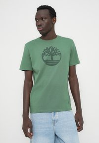 Πράσινο βαμβακερό μπλουζάκι με μαύρο λογότυπο δέντρου στο μπροστινό μέρος, στρογγυλή λαιμόκοψη και κοντά μανίκια. Συνδυάζεται με ανοιχτό μπλε τζιν σορτς.