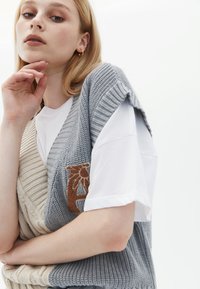 Gilet lavorato a maglia in grigio chiaro e crema, con texture a coste e una toppa floreale marrone. Sotto, una t-shirt bianca a maniche corte.