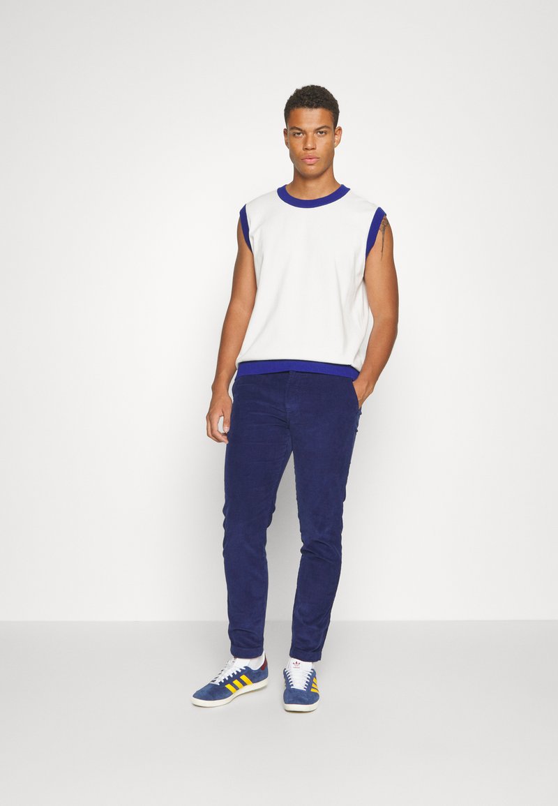 Homme debout les mains dans les poches, portant un pull sans manches blanc avec des bordures bleues, un pantalon bleu et des baskets bleues avec des rayures jaunes.