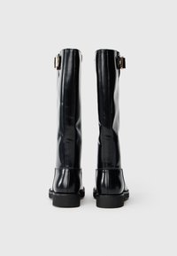 Botas negras hasta la rodilla, hechas de un material brillante con una textura suave, que cuentan con una punta redondeada y un acento de hebilla dorada en el costado.