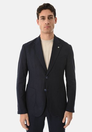 Uomo con capelli corti e scuri che indossa un blazer blu navy scuro sopra un maglione a coste beige, in piedi contro uno sfondo chiaro e uniforme.