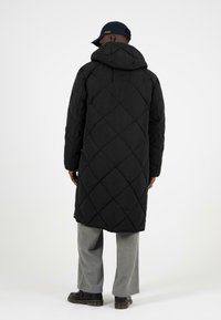 Černý prošívaný dlouhý kabát s kapuc�í, zdobený diamantovými vzory; nošený přes světle šedé kalhoty a černé boty, pohled zezadu.