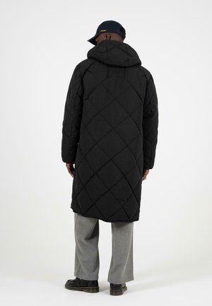 Cappotto lungo trapuntato nero con cappuccio, caratterizzato da motivi a rombi; indossato sopra pantaloni grigio chiaro e scarpe nere, visto da dietro.