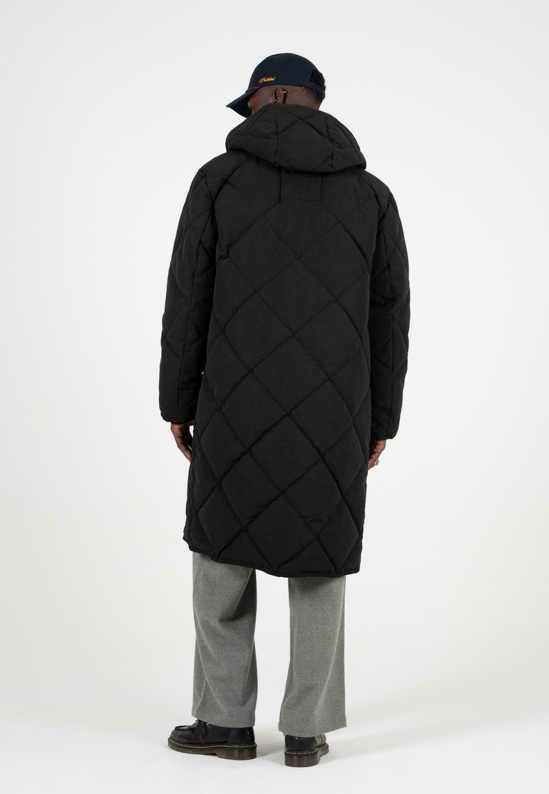 Černý prošívaný dlouhý kabát s kapucí, zdobený diamantovými vzory; nošený přes světle šedé kalhoty a černé boty, pohled zezadu.