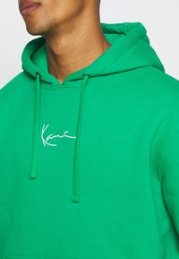 Uomo che indossa una felpa verde con cappuccio e lacci, con un logo in stile firma bianco sul petto, tagliata all'altezza del mento e della spalla.
