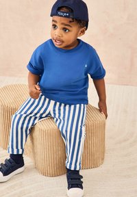Camiseta azul de manga corta, pantalones a rayas blancos y azules, zapatillas navy y una gorra navy, sentado en un taburete redondo texturizado en color beige.