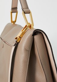 Tan leather handbag with gold hardware, featuring a structured design, visible stitching, and contrasting edges.