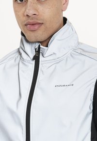 Silberne und schwarze leichte Jacke mit Reißverschluss auf der Vorderseite, hohem Kragen und Logo auf der linken Brust. Glatte Textur und taillierte Passform.