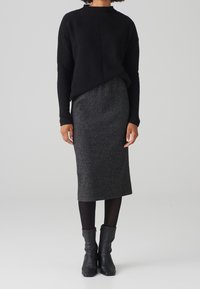 Personne portant un pull noir, une jupe grise foncée au genou, des collants noirs et des bottines noires, debout contre un fond clair et uni.