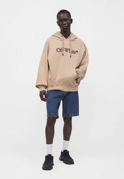Jeune homme portant un sweat à capuche beige Off-White, un short en jean bleu, des chaussettes blanches et des baskets noires, debout devant un fond blanc uni.