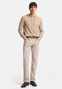 Maglione beige con cappuccio realizzato in tessuto morbido, abbinato a pantaloni beige chiaro e scarpe in pelle marrone scuro, con una cintura semplice.