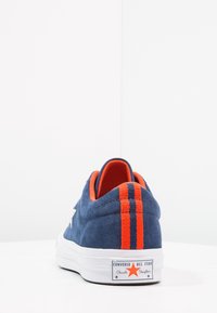 Zapatilla de ante azul marino con un interior naranja y dos rayas verticales en el talón. Suela de goma blanca con etiqueta de marca en el lateral.
