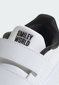 White leather sneaker with black inner lining, featuring a sturdy strap and embossed "Smiley World" logo on the heel. Textured details present.