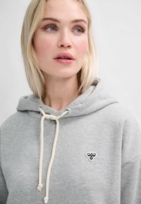 Blonde Frau trägt einen hellgrauen Hoodie mit weißen Kordeln und einem kleinen schwarz-weißen Bienen-Emblem auf der Brust, blickt nach rechts.