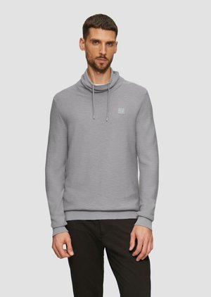 Lichtgrijze sweatshirt met een opgerolde kraag, koordjes en logo patch. De stof heeft een gestructureerde afwerking, gecombineerd met donkere broek.