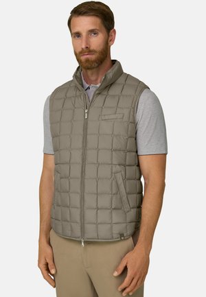 Mann trägt eine hellbraune gesteppte ärmellose Weste über einem grauen kurzärmligen Poloshirt und beigen Hosen, steht mit neutralem Gesichtsausdruck.
